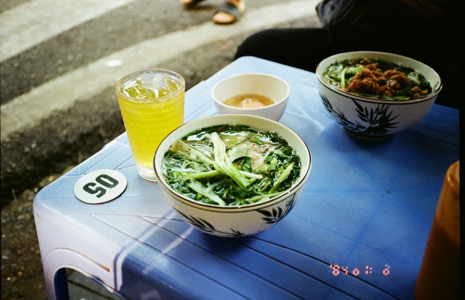 เฝอในชามลายไม้ไผ่กับชาเขียวเย็นบนโต๊ะพลาสติกสีฟ้าริมถนนในฮานอย